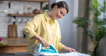 Waist-up portrait of a cheerful woman with an iron in one hand leaning over the ironing-board