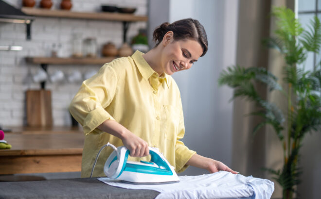 Waist-up portrait of a cheerful woman with an iron in one hand leaning over the ironing-board