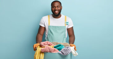 Delighted satisfied plump black man rejoices finishing washing day successfully and in time, holds plastic container with clean fresh laundry, stands against blue wall, maintances housekeeping