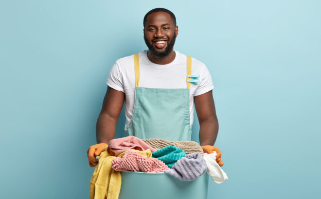 Delighted satisfied plump black man rejoices finishing washing day successfully and in time, holds plastic container with clean fresh laundry, stands against blue wall, maintances housekeeping